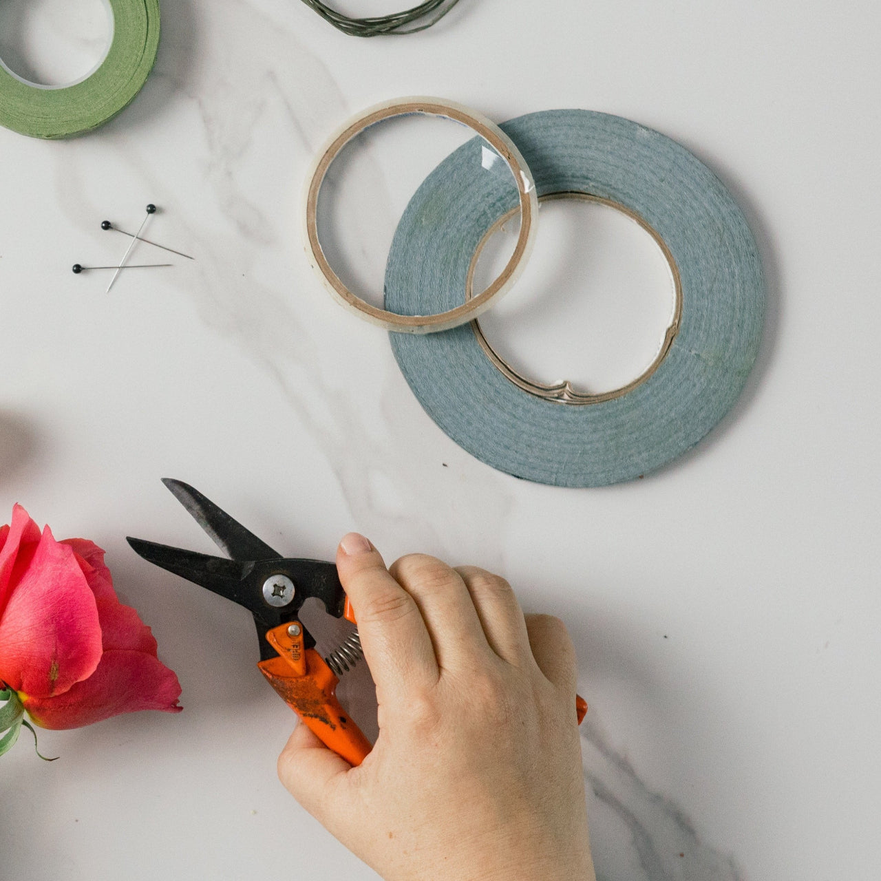 A person is cutting a rose with scissors, while various crafting materials like wire, fabric, and beads are laid out on a white surface.