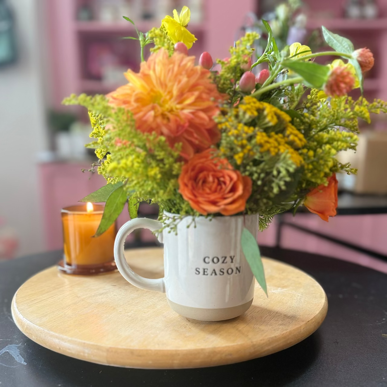 A floral arrangement in a white ceramic mug with the words 'COZY SEASON' printed on it, placed on a round wooden tray on a black table. The bouquet includes orange flowers, yellow wildflowers, and green foliage, with a small amber candle in the background.