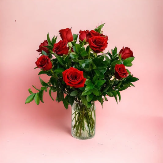 A dozen red roses in a clear glass vase against a pink background.
