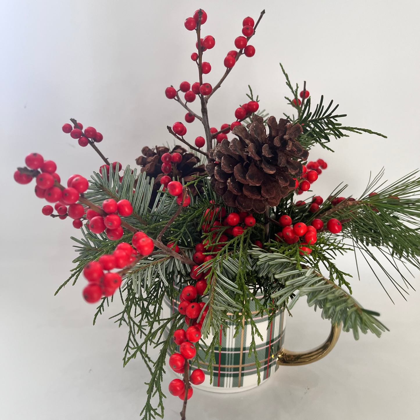Decorative arrangement with red berries, pine cones, and greenery in a plaid mug on a white background