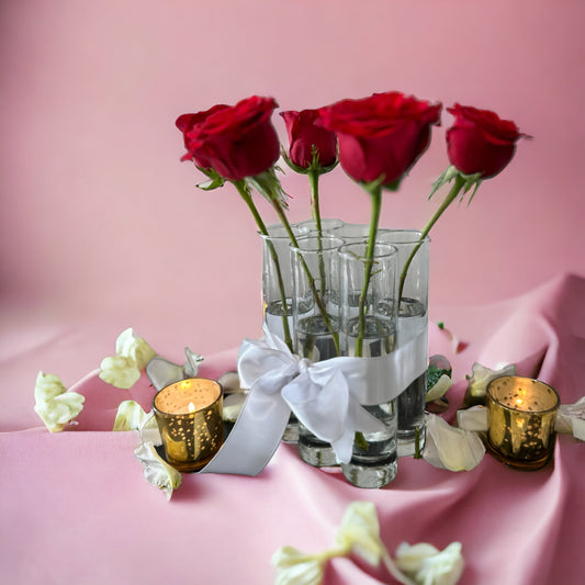 A set of six red roses placed in clear glass bud vases, tied with white satin ribbons, set against a pink background with decorative gold elements.