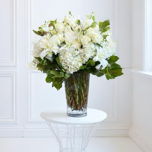 Bouquet of white flowers in a clear vase on a white pedestal against a white wall.
