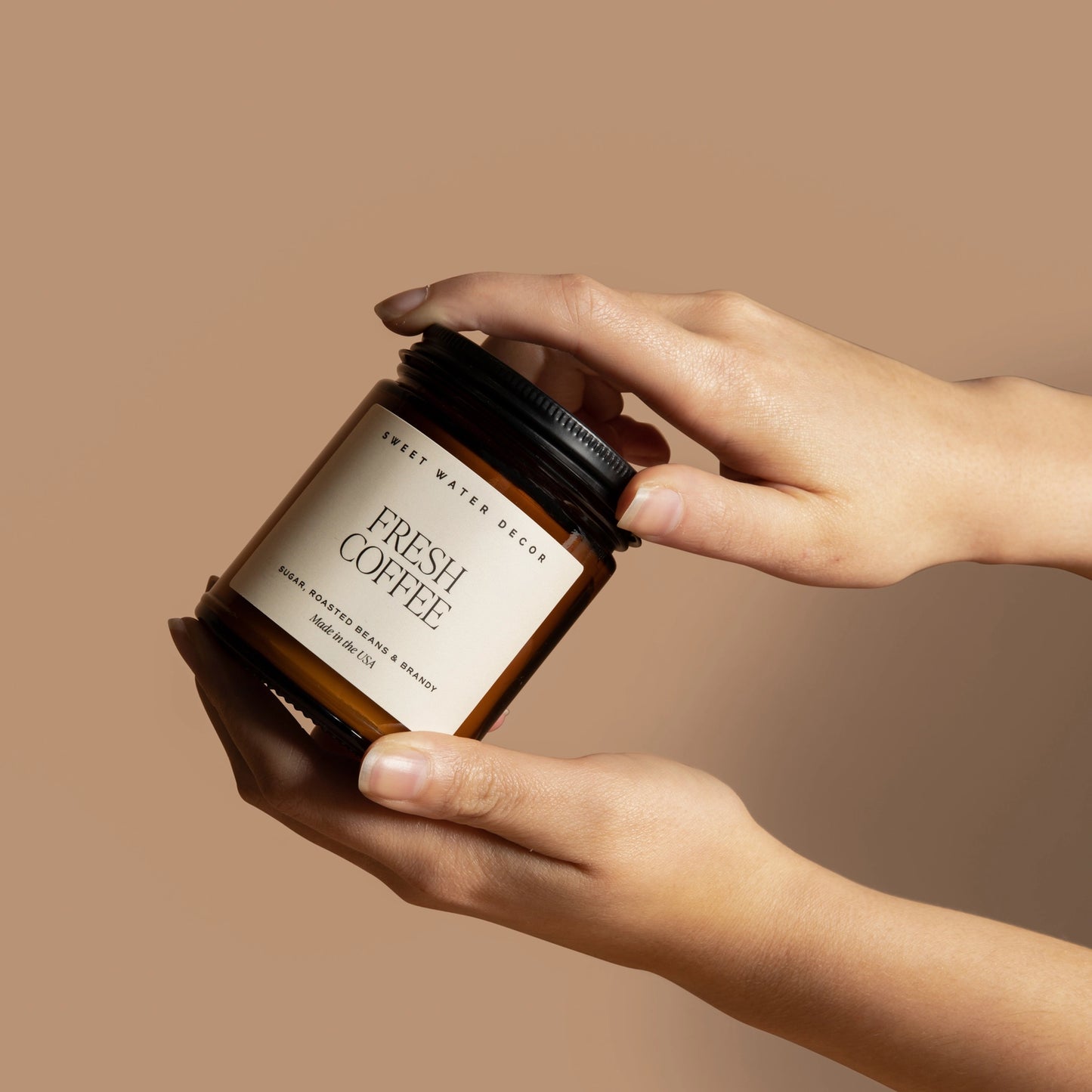 Hand holding a candle labeled 'Fresh Coffee' against a beige background