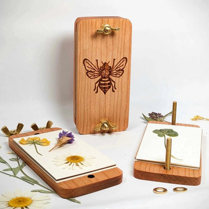 A wooden pocket flower press with a hand-drawn bee design, displayed alongside pressed flowers and adjustable brass screws.