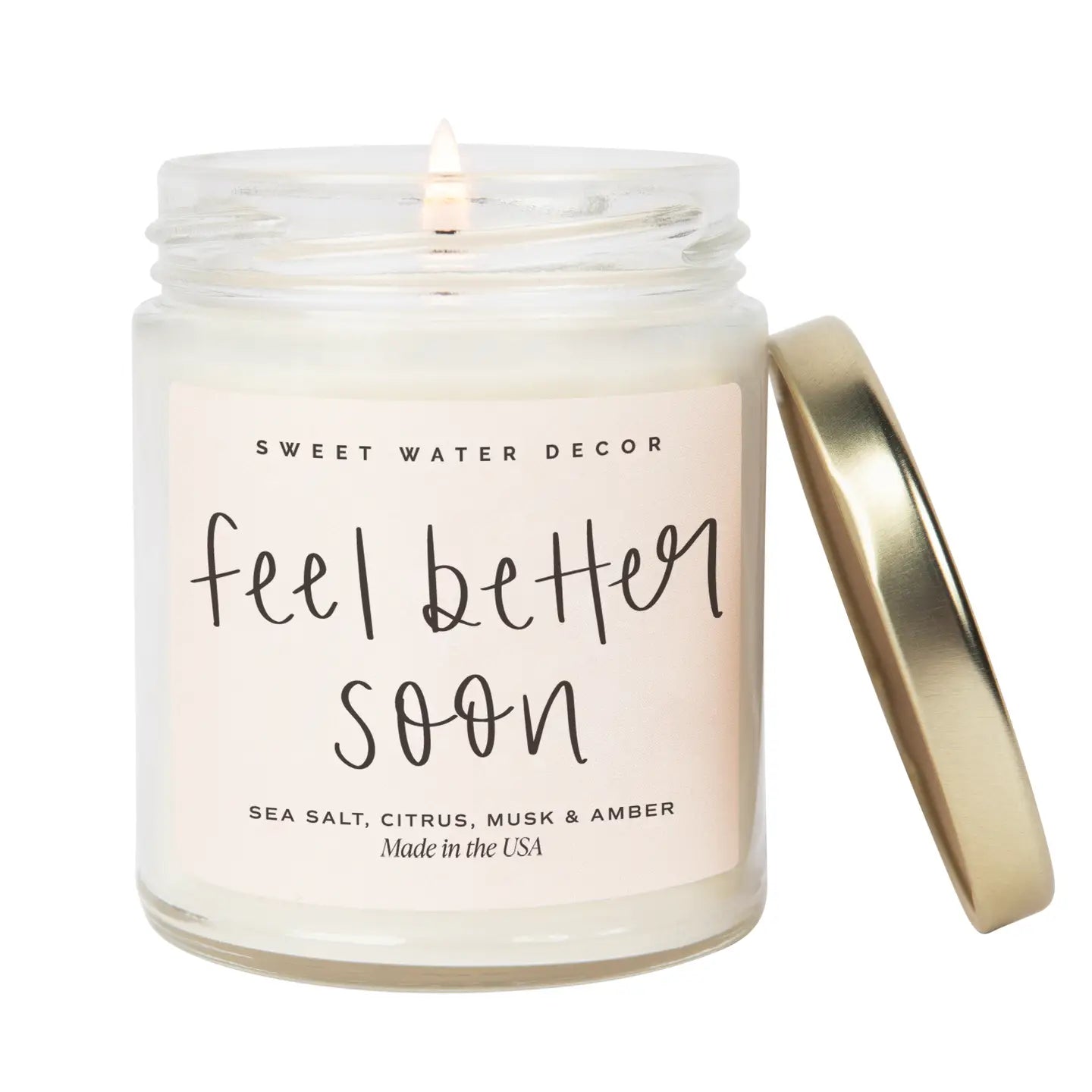 A clear glass jar soy candle with a pink label that reads 'Feel Better Soon' along with the scent notes of sea salt, citrus, musk, and amber. The candle is closed with a metallic lid and is placed on a white surface.