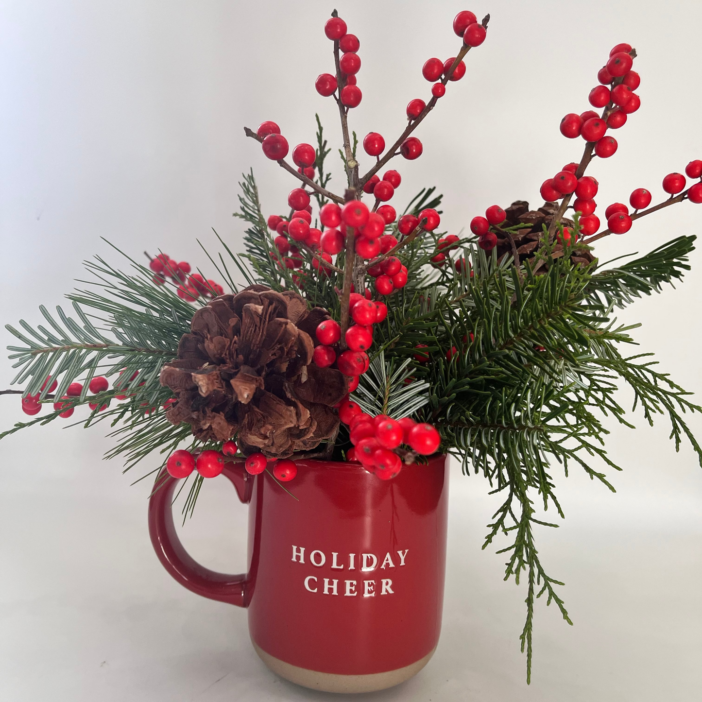 Red mug with 'Holiday Cheer' text, filled with greenery and red berries on a white background