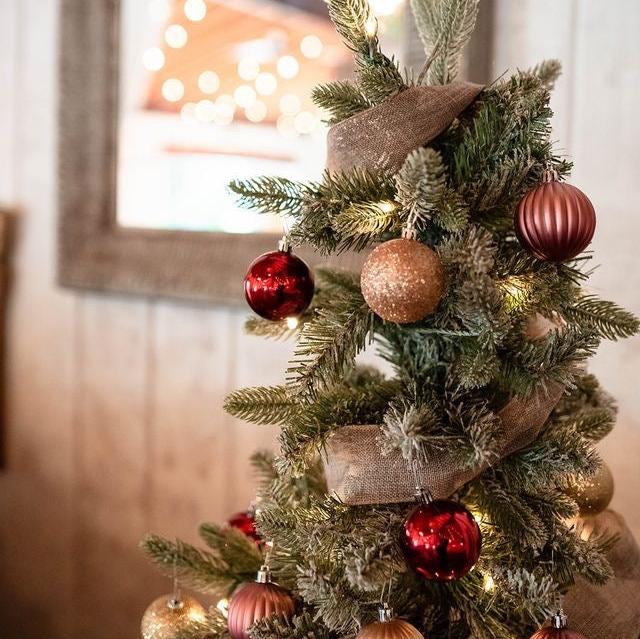 A miniature Christmas tree centerpiece with artificial green branches, brown trimmings, and red and gold ornaments, set against a rustic wooden backdrop with Christmas lights.
