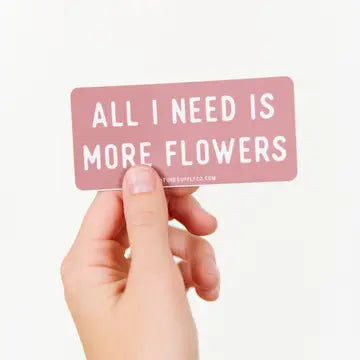 A hand holding a pink vinyl sticker with the text 'ALL I NEED IS MORE FLOWERS'.