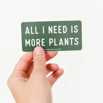 A hand holding a green vinyl sticker with the text 'All I need is more plants'.