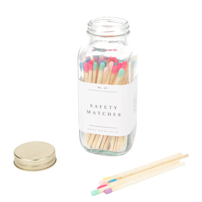 A glass jar containing multicolored safety matches with a label on the front that reads 'Safety Matches'. There are a few matches displayed outside of the jar, and a metal lid is also shown to the side.