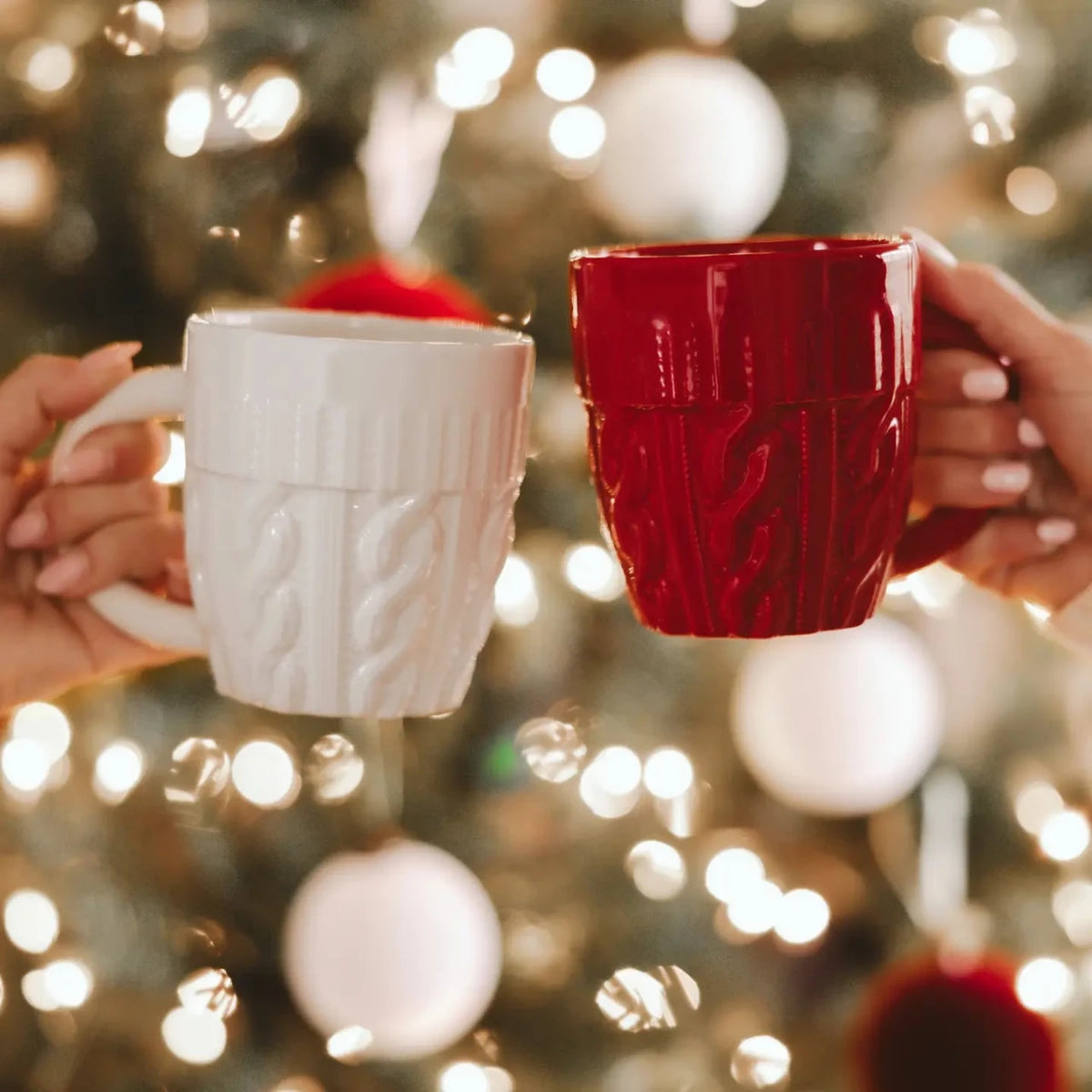Two hands holding white and red textured mugs in front of a blurred Christmas tree with lights.