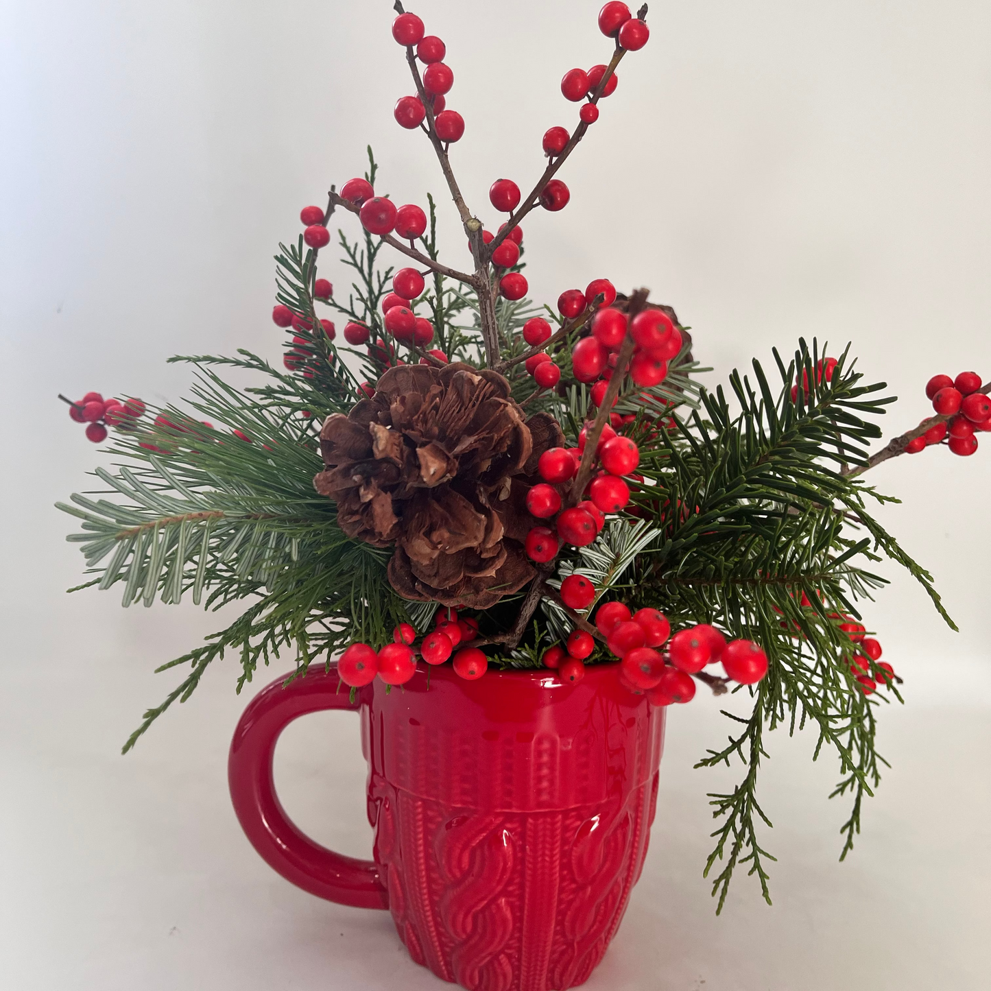 Red mug with festive arrangement of greenery, red berries, and pine cones on a white background
