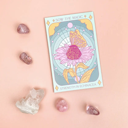 A tarot card with a floral design representing the 'Strength' tarot card, alongside a pink crystal, a white crystal, and a pink agate gemstone on a pink background.