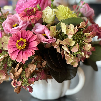 A floral arrangement in a white ceramic pumpkin-shaped mug, with a variety of flowers including pink and green, and autumnal tones.