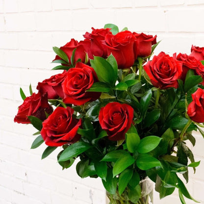 Two dozen long-stemmed roses are accented with seasonal foliage in an elegant glass vase.