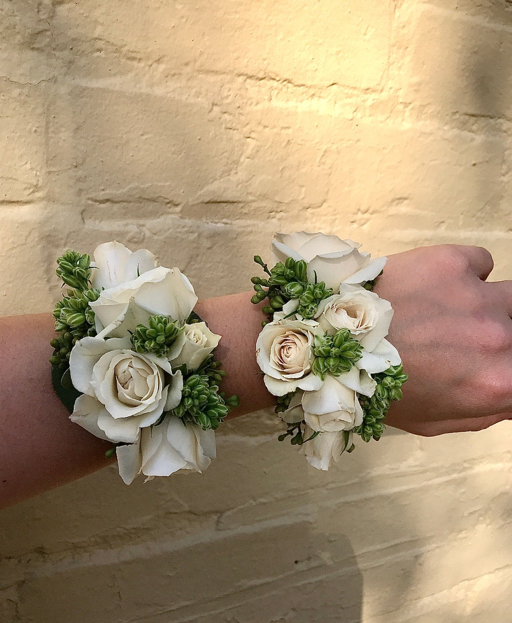 Two wrist corsages made from spray roses and texture, against a yellow brick wall. 