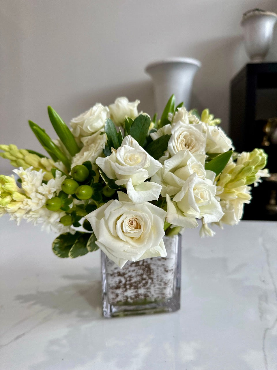 Wintry white plumes of fragrant hyacinth are paired with snowflake spray roses and green hypericum berries. Decorated simply with variegated foliage in a white birch-lined vase